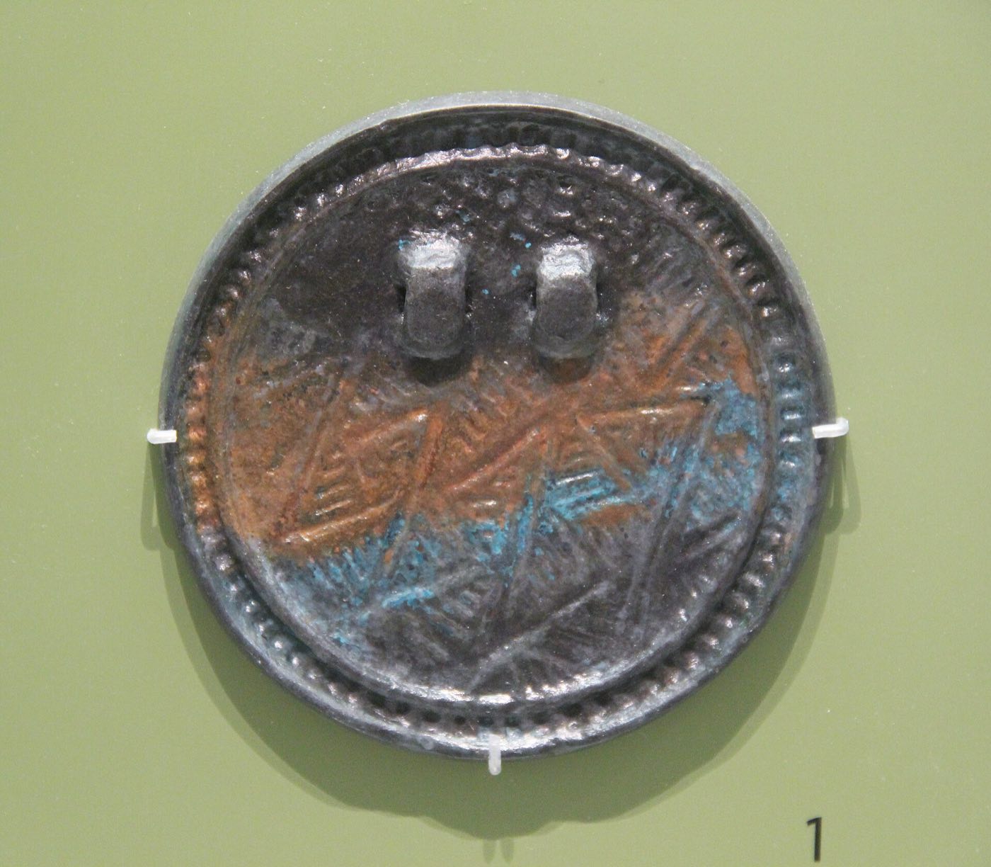 Cast bronze mirror with a coarse zigzag pattern and two loops on the back. The loops held a cord, so mirrors like this could be hung on the body as a sign of a ritual leader.