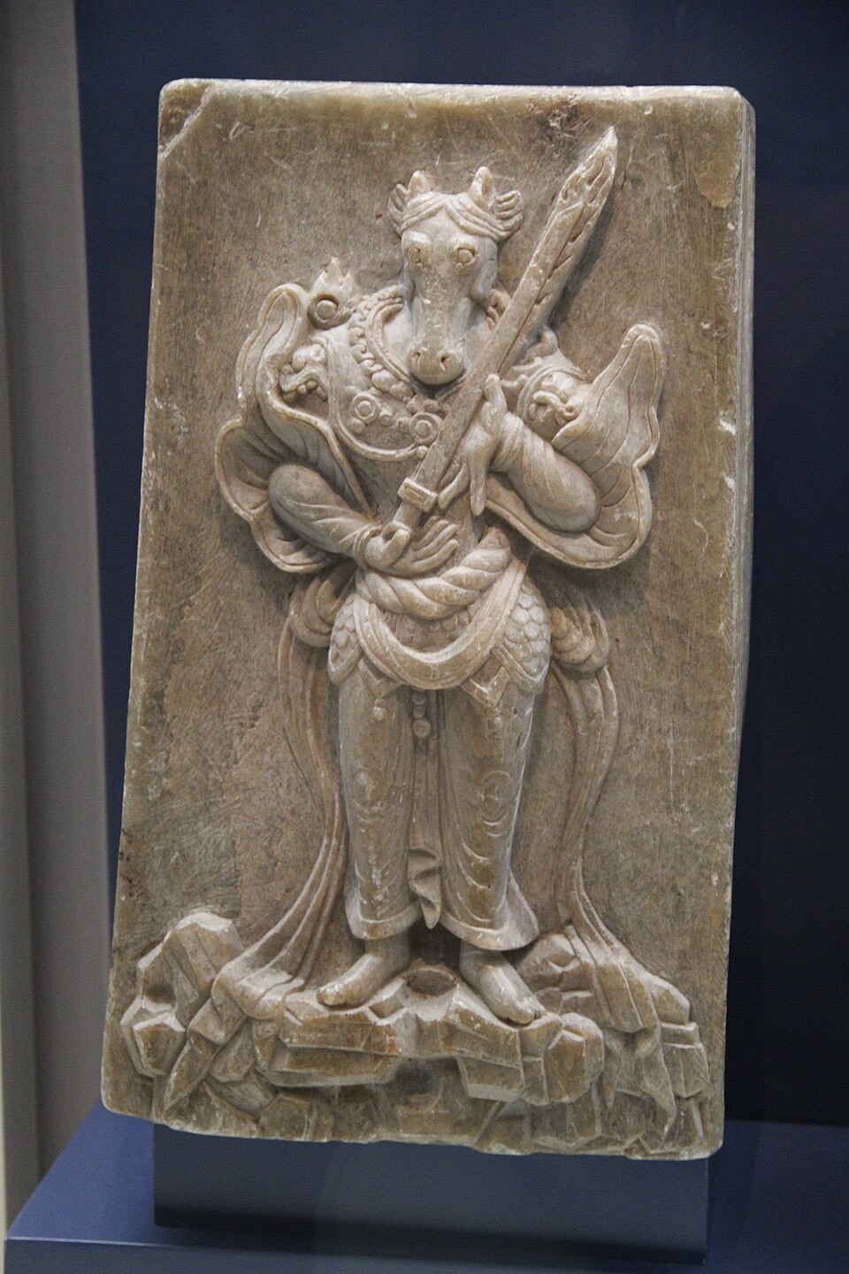 A guardian carved on a stone panel, of the kind set around Unified Silla royal tombs. He wears scale armour to the knee, a skirt of plates, shoulder guards and a crested helm, and holds a weapon upright. It is the closest thing to a portrait of a Silla soldier in his kit.